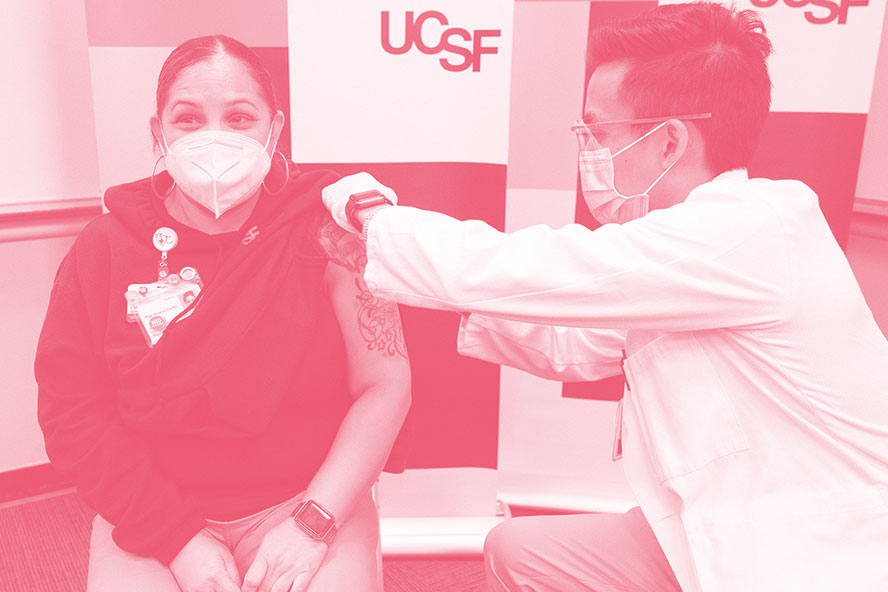 Reina Lopez, wearing a mask, sits with her sleeve rolled up as a U C S F doctor administers her vaccine shot