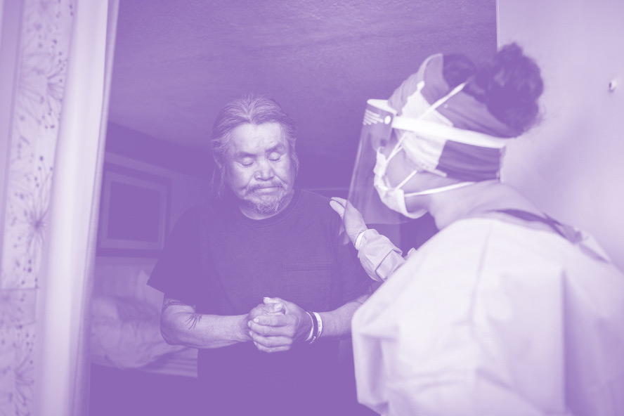 A Navajo man, hands folded and eyes closed, is comforted by a U C S F doctor wearing P P E gear